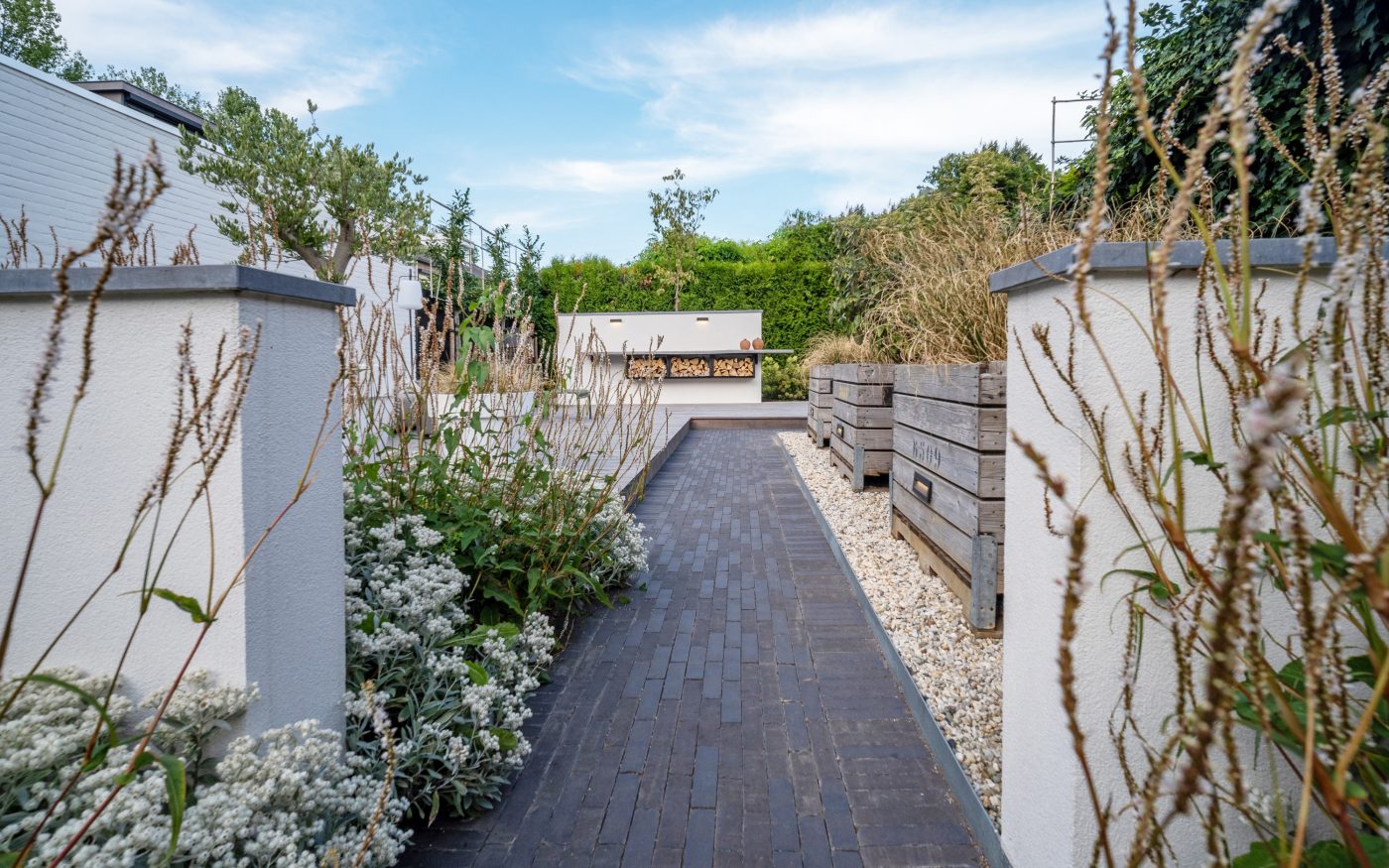 Een pad van donkere gebakken klinkers tussen beplanting en witte tuinmuren